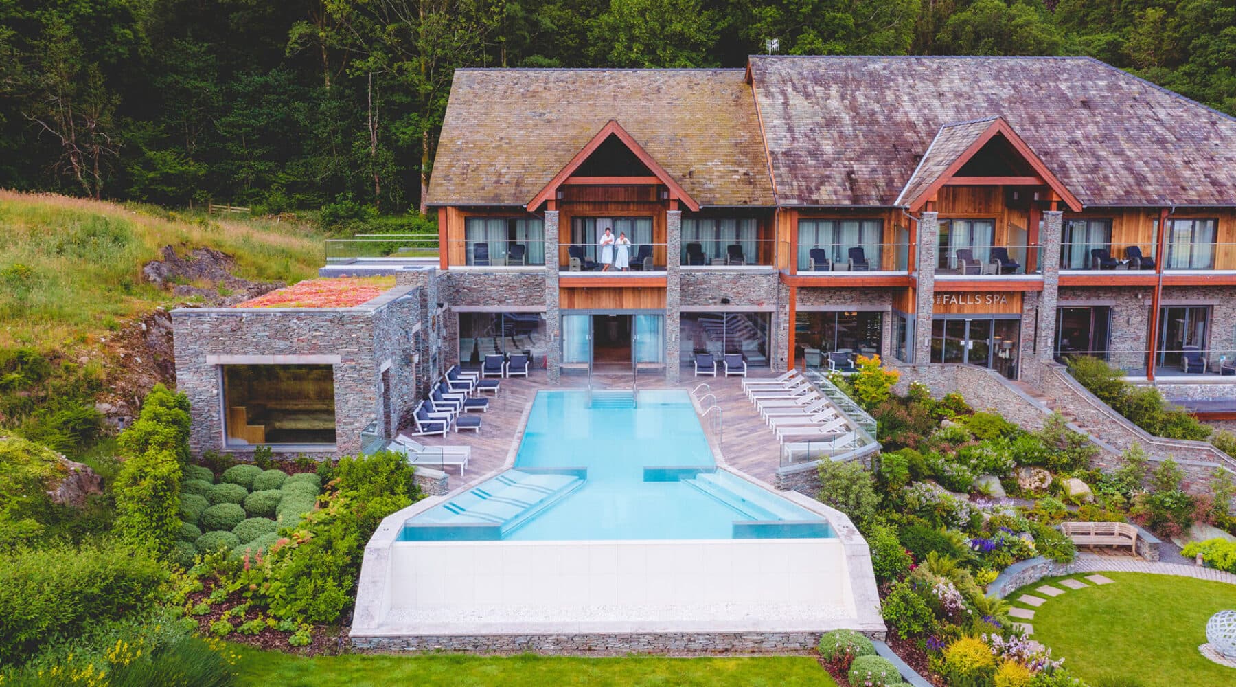 Stunning aerial view of The Falls Spa at Lodore Falls Hotel in Borrowdale showing multi-level outdoor hydrotherapy pools, traditional Lakeland slate and stone architecture, manicured gardens with sun loungers, and dramatic Lake District mountain backdrop, showcasing the complete spa resort facilities