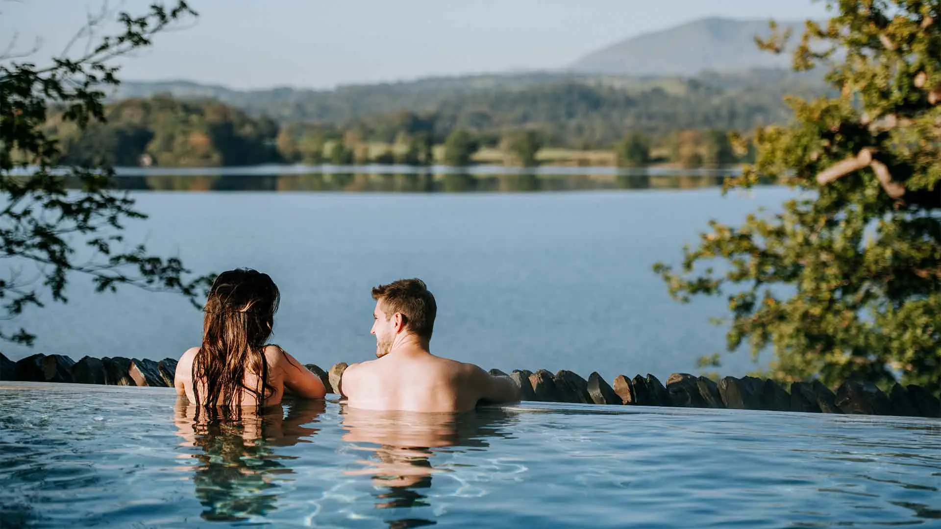 Romantic couple enjoying stunning infinity-edge vitality pool at Low Wood Bay Resort and Spa, gazing across Lake Windermere with panoramic Lake District mountain and woodland views, perfect for couples spa breaks and romantic getaways in Cumbria