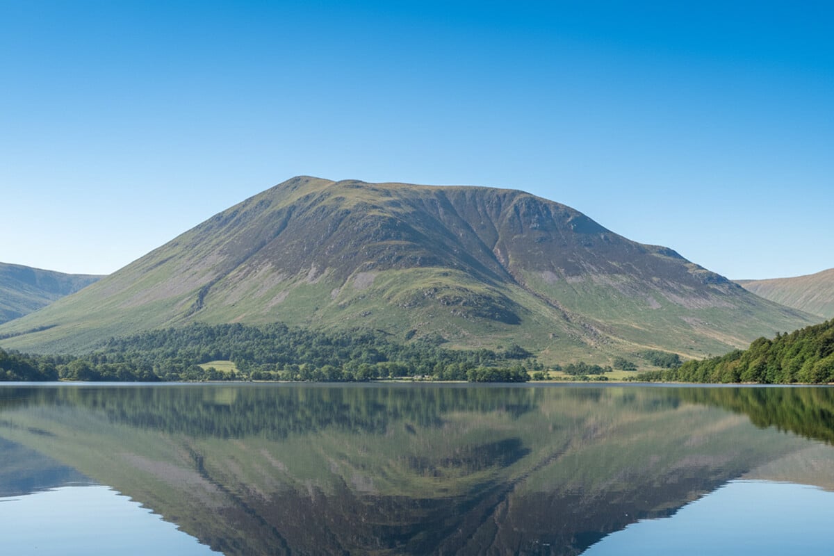 Dramatic fell reflected in the mirror-still waters of Bassenthwaite Lake on a clear summer day, with native woodland along the shoreline in the northern Lake District near Keswick