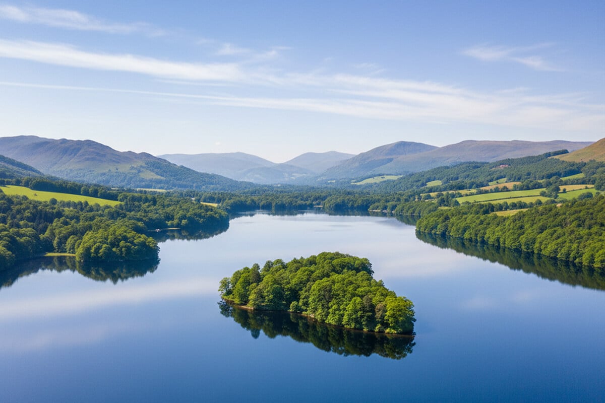 Aerial view of a tree-covered island surrounded by calm blue water on Lake Windermere near Bowness, with wooded shores and rolling Lakeland fells stretching into the distance on a summer morning