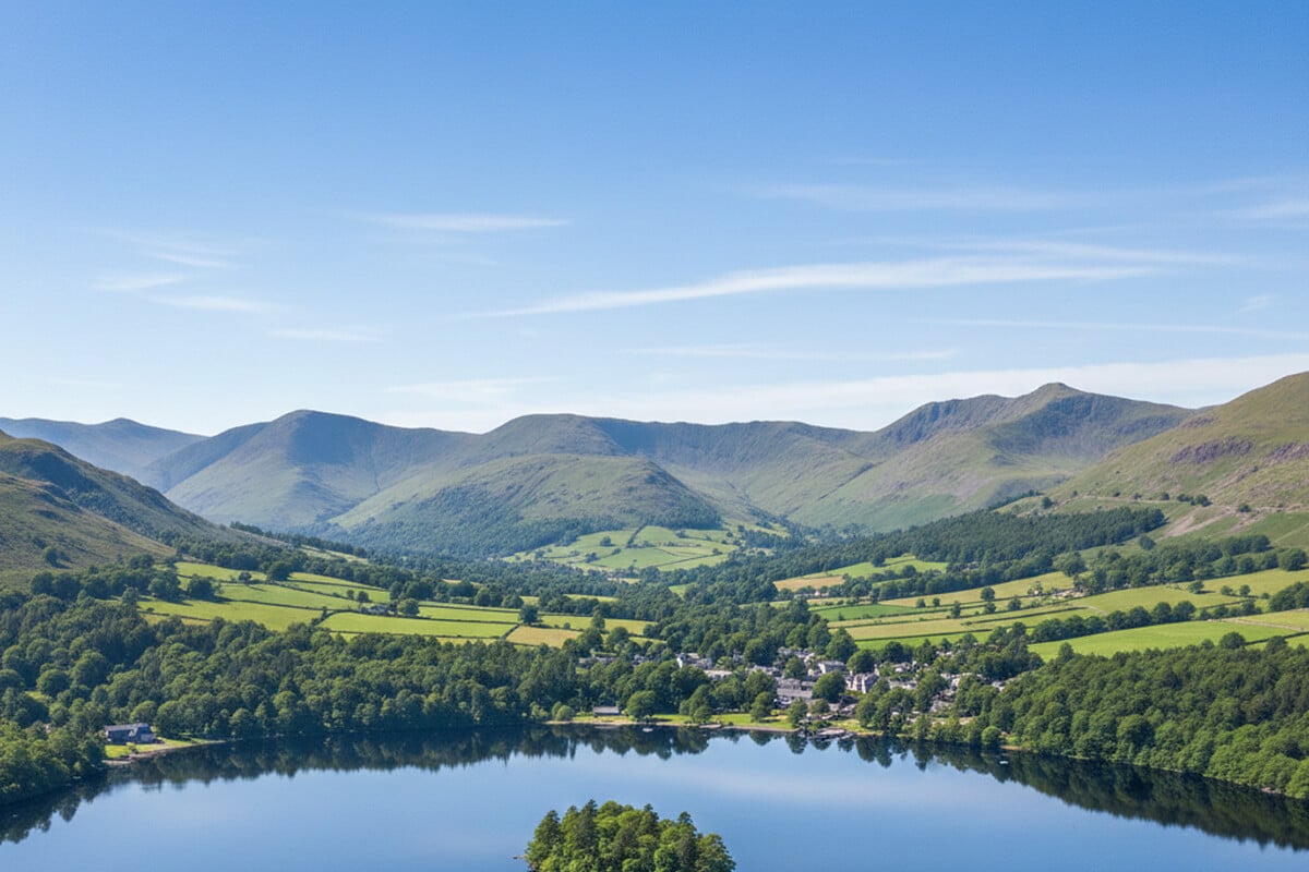 Aerial view of Grasmere village beside its lake with a tree-covered island in the foreground, green fields and mature woodland leading up to rounded Lakeland fells under a clear summer sky in the Lake District