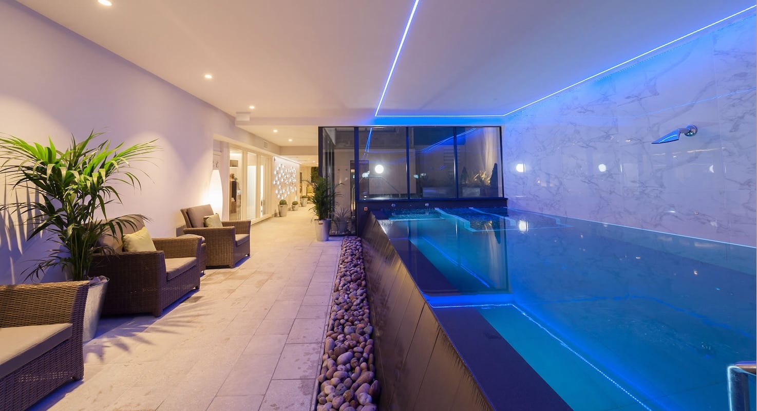 Contemporary hydrotherapy pool at Appleby Manor Country House Hotel near Appleby-in-Westmorland, featuring dramatic blue LED underwater lighting, natural Lake District pebble stone border detail, modern wicker relaxation seating with tropical palms, and luxury spa facilities in the Eden Valley