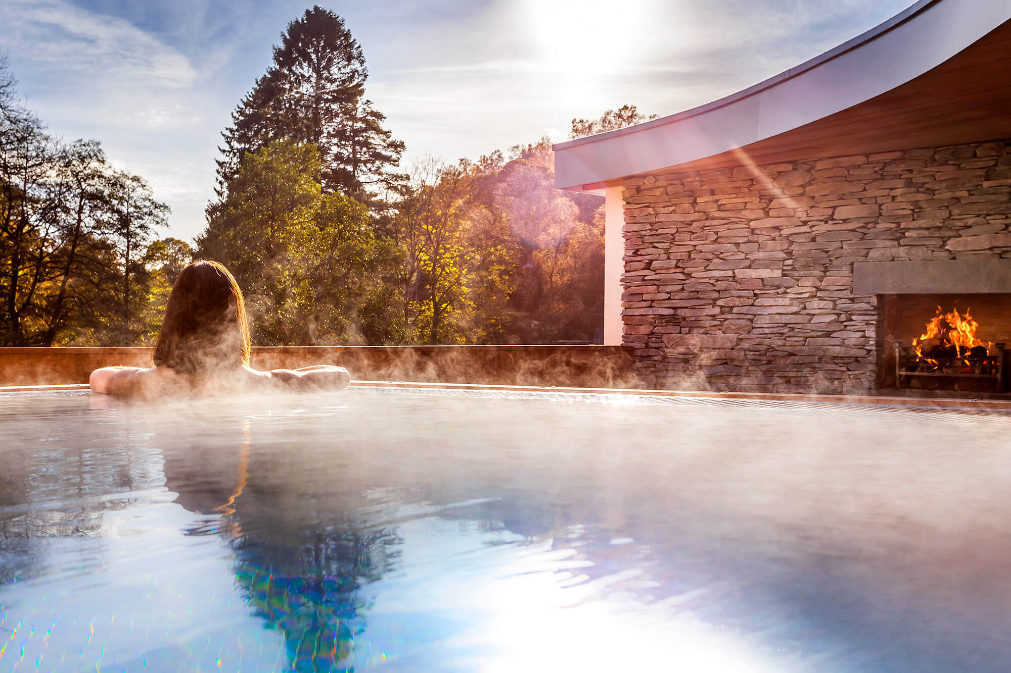 Guest enjoying steaming outdoor hydrotherapy pool at Brimstone Hotel and Spa in Langdale at golden hour sunset, with Lakeland dry stone fireplace, thermal mist rising, and Lake District woodland creating magical Nordic spa experience in Cumbria