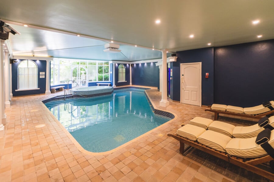 Elegant indoor heated pool at The Grange Boutique Hotel in Grange-over-Sands featuring sophisticated navy blue accent walls, classic terracotta tile flooring, contemporary wooden loungers with cream cushions, floor-to-ceiling windows with garden views, Victorian-era grandeur meets modern wellness on Morecambe Bay