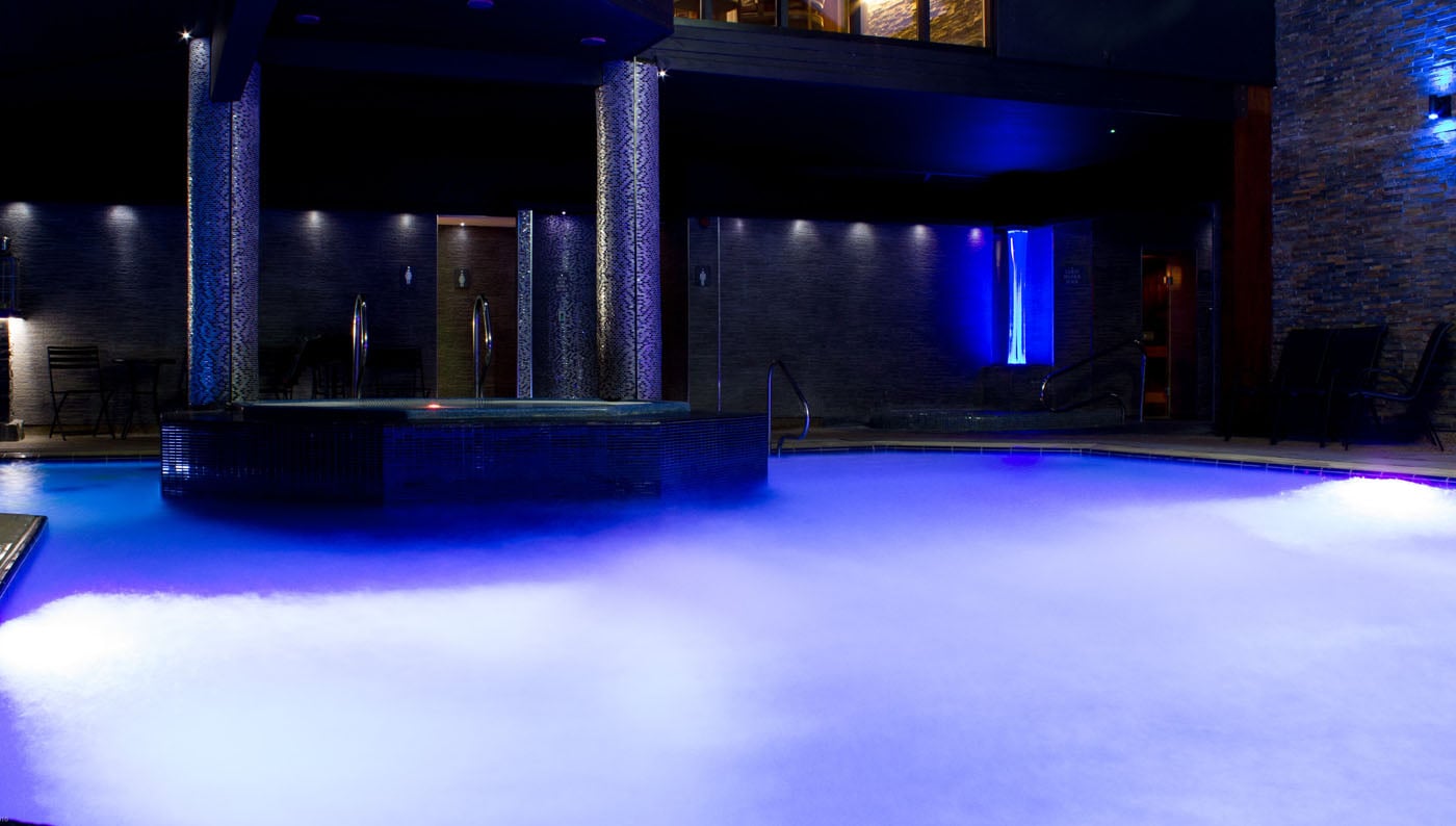Atmospheric main swimming pool at Lakes Hotel and Spa in Bowness-on-Windermere at dusk featuring dramatic blue LED mood lighting, contemporary iridescent mosaic architectural columns, signature bubble pool with secret button feature, extended evening hours creating moody Lake District spa experience