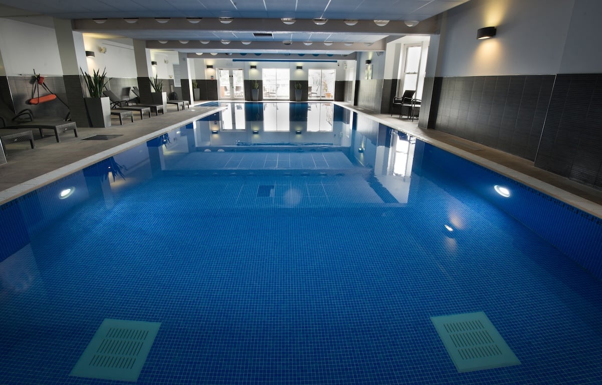 Modern indoor swimming pool at Macdonald Old England Hotel and Spa in Bowness-on-Windermere, featuring deep blue mosaic tiling, contemporary black and white interior design, underwater lighting, and heated pool facilities in a luxurious Lake District spa setting