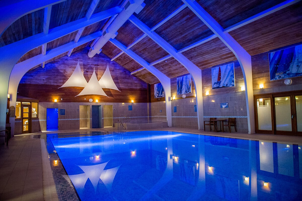 Spectacular 13-metre indoor pool at North Lakes Hotel and Spa in Penrith featuring dramatic blue LED lighting, vaulted timber ceiling with exposed architectural beams creating cathedral-like atmosphere, contemporary white pendant lights, family-friendly swimming facilities near Ullswater in the Lake District