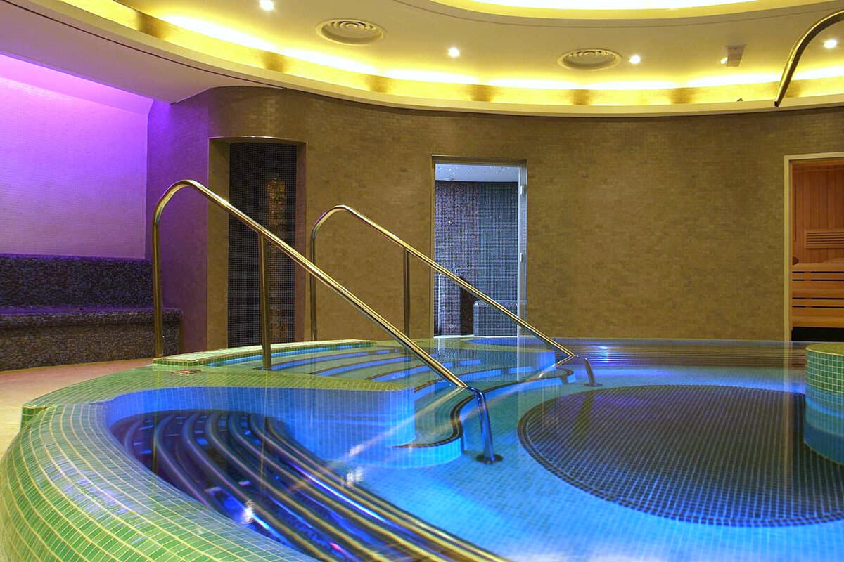Multi-level hydrotherapy vitality pool at Armathwaite Hall Hotel and Spa near Bassenthwaite Lake, featuring dramatic blue LED underwater lighting with green gradient mosaic tiles, curved architectural design with chrome handrails, golden ambient ceiling lighting and purple accent walls, leading to Finnish sauna and thermal suite facilities in award-winning Lake District spa Armathwaite Hall Hotel & Spa