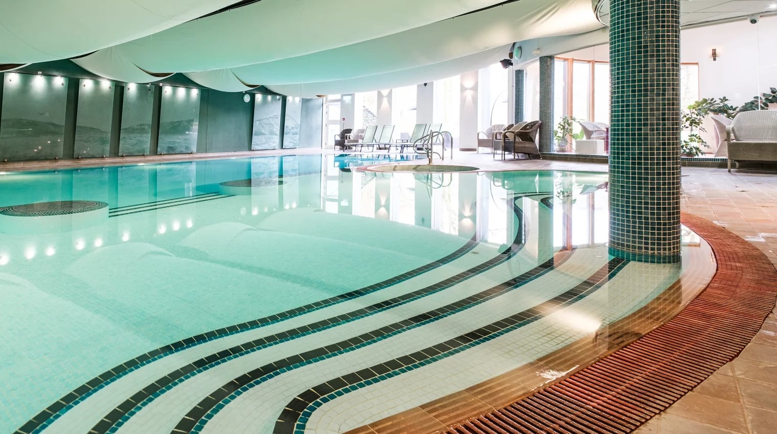 Contemporary indoor pool at Lakeside Hotel Health Club and Spa on Lake Windermere, featuring distinctive teal and green mosaic tile columns, flowing wave-pattern ceiling design, and spacious swimming area with loungers overlooking the Lake District waterfront