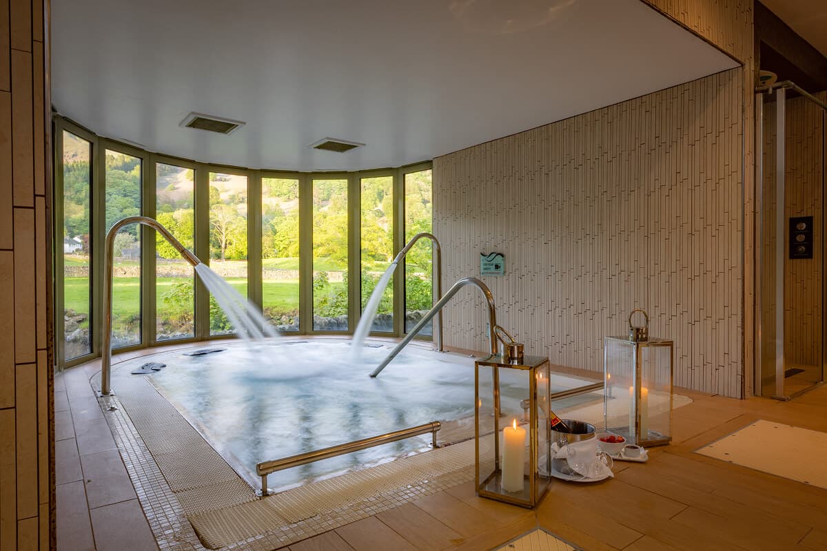 HydroSpa hydrotherapy pool at Rothay Garden by Harbour Hotels in Grasmere village, featuring powerful cascade waterfall features into circular hot tub with sunken loungers, bamboo-textured walls, and expansive fell views through floor-to-ceiling windows in the heart of the Lake District