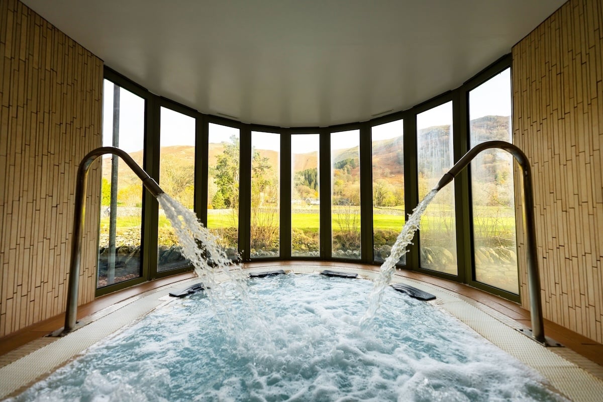 Panoramic hydrotherapy pool at Rothay Garden Hotel and Riverside Spa in Grasmere, featuring curved floor-to-ceiling windows with dramatic Lake District fell views, dual cascade water features, bamboo-panelled walls, and intimate circular hot tub design surrounded by nature