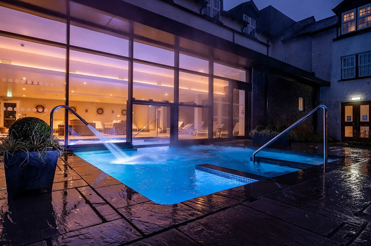 Relaxing in heated outdoor hydrotherapy pool at Swan Hotel and Spa Newby Bridge, guest enjoying cocktail in naturally filtered swim-through pool with minimal chlorine, contemporary adults-only spa atmosphere with white loungers and wooden decking on River Leven near Lake Windermere in Cumbria