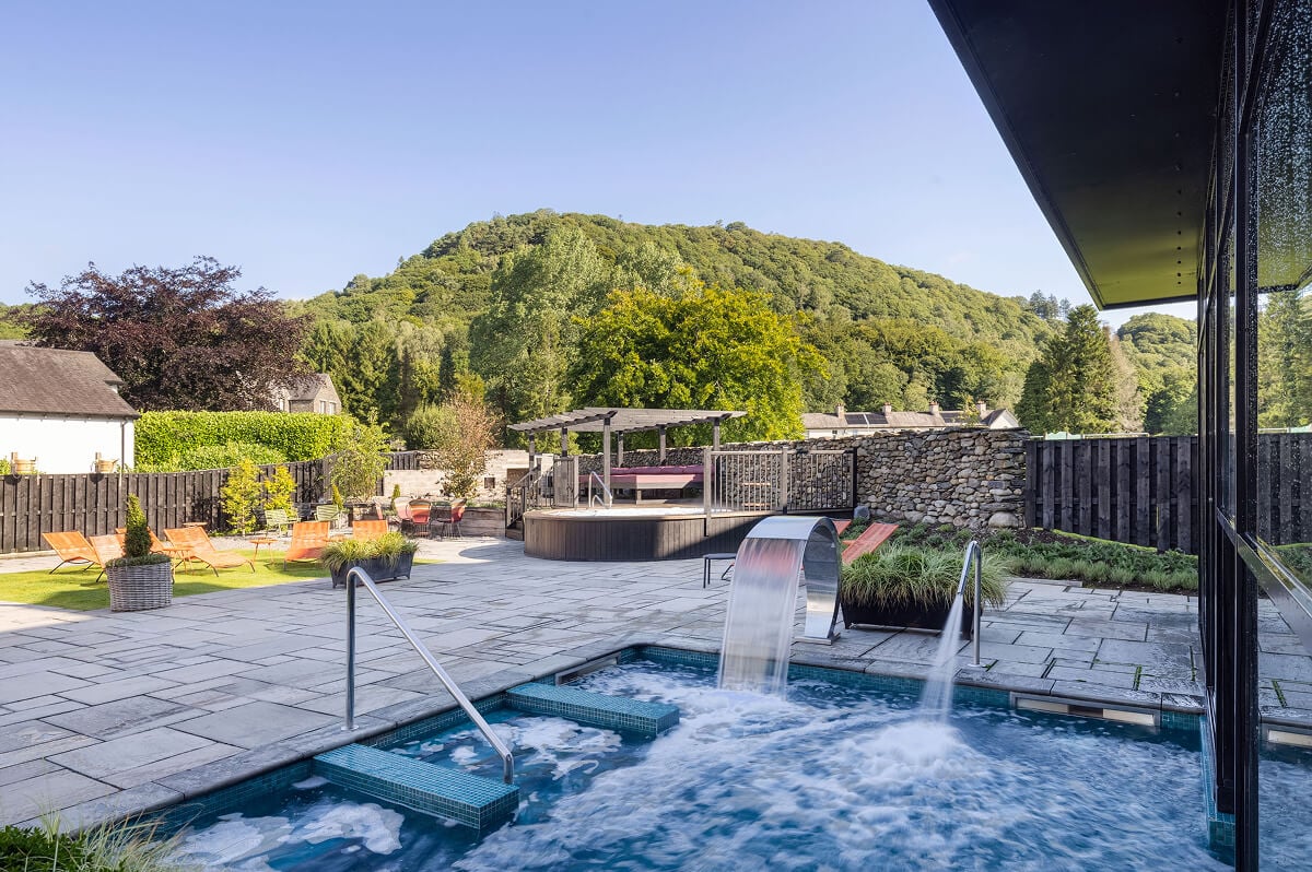 Outdoor thermal spa garden at Swan Hotel Holte Spa in Newby Bridge featuring bubbling hydrotherapy pool with dramatic cascade waterfall features, wooden hot tub with dramatic Lake District fell views, spacious decking with loungers, and authentic Finnish sauna overlooking River Leven in Cumbria