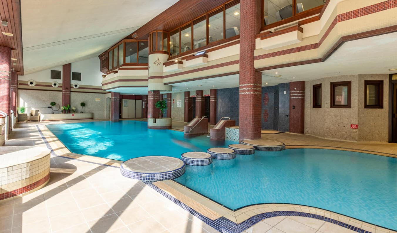 Expansive leisure pool at Whitewater Hotel Country Club near Newby Bridge, featuring multi-level design with mosaic-tiled stepping stones, burgundy and cream decorative columns, spacious swimming area, and grand architectural poolside complex in the Lake District Whitewater Hotel